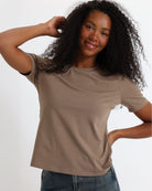 A woman wearing a brown short-sleeve crewneck t-shirt and jeans, standing against a fabric backdrop.