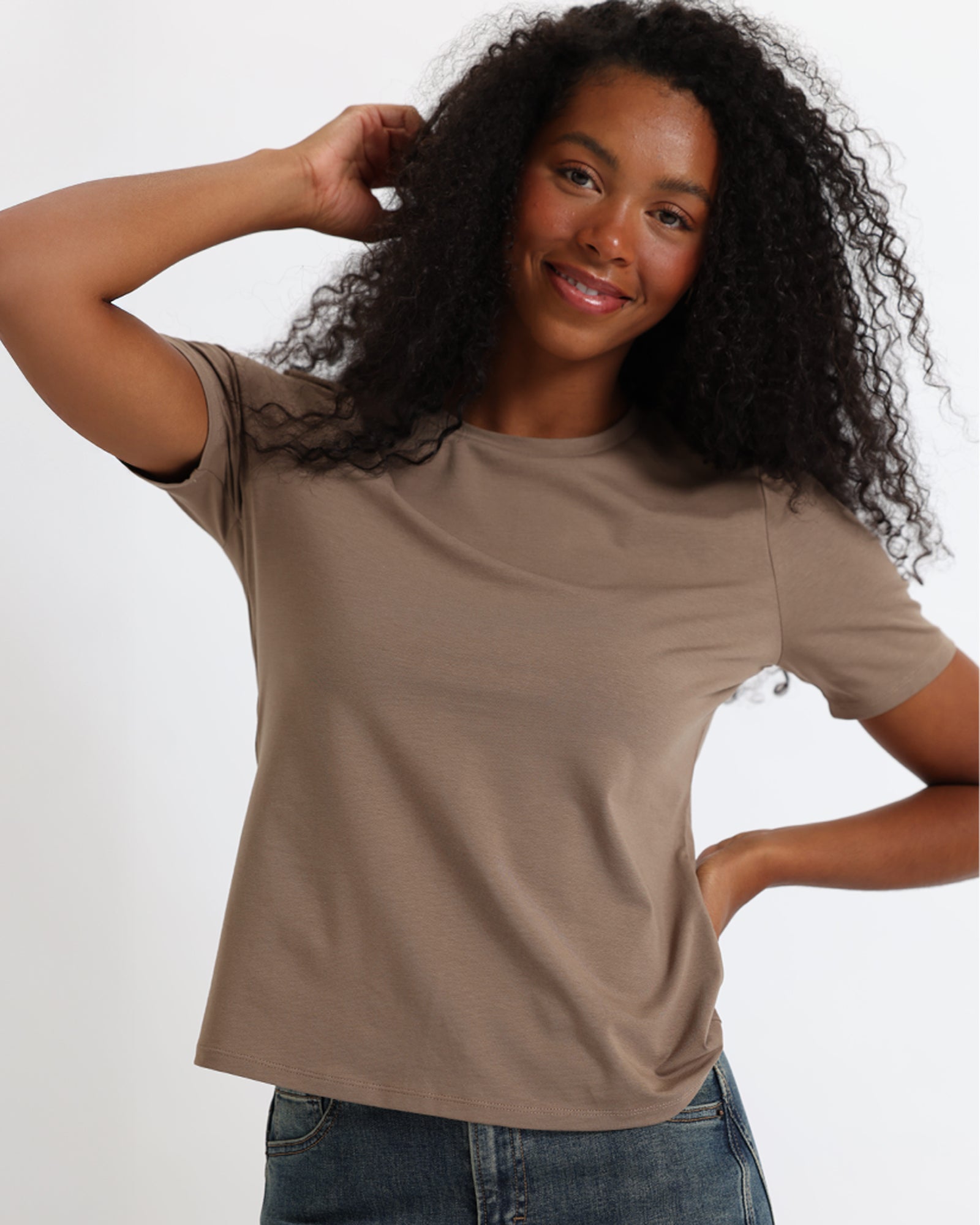 A woman wearing a brown short-sleeve crewneck t-shirt and jeans, standing against a fabric backdrop.