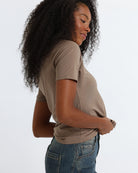 A woman wearing a brown short-sleeve crewneck t-shirt and jeans, standing against a fabric backdrop.