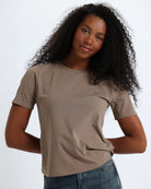 A woman wearing a brown short-sleeve crewneck t-shirt and jeans, standing against a fabric backdrop.