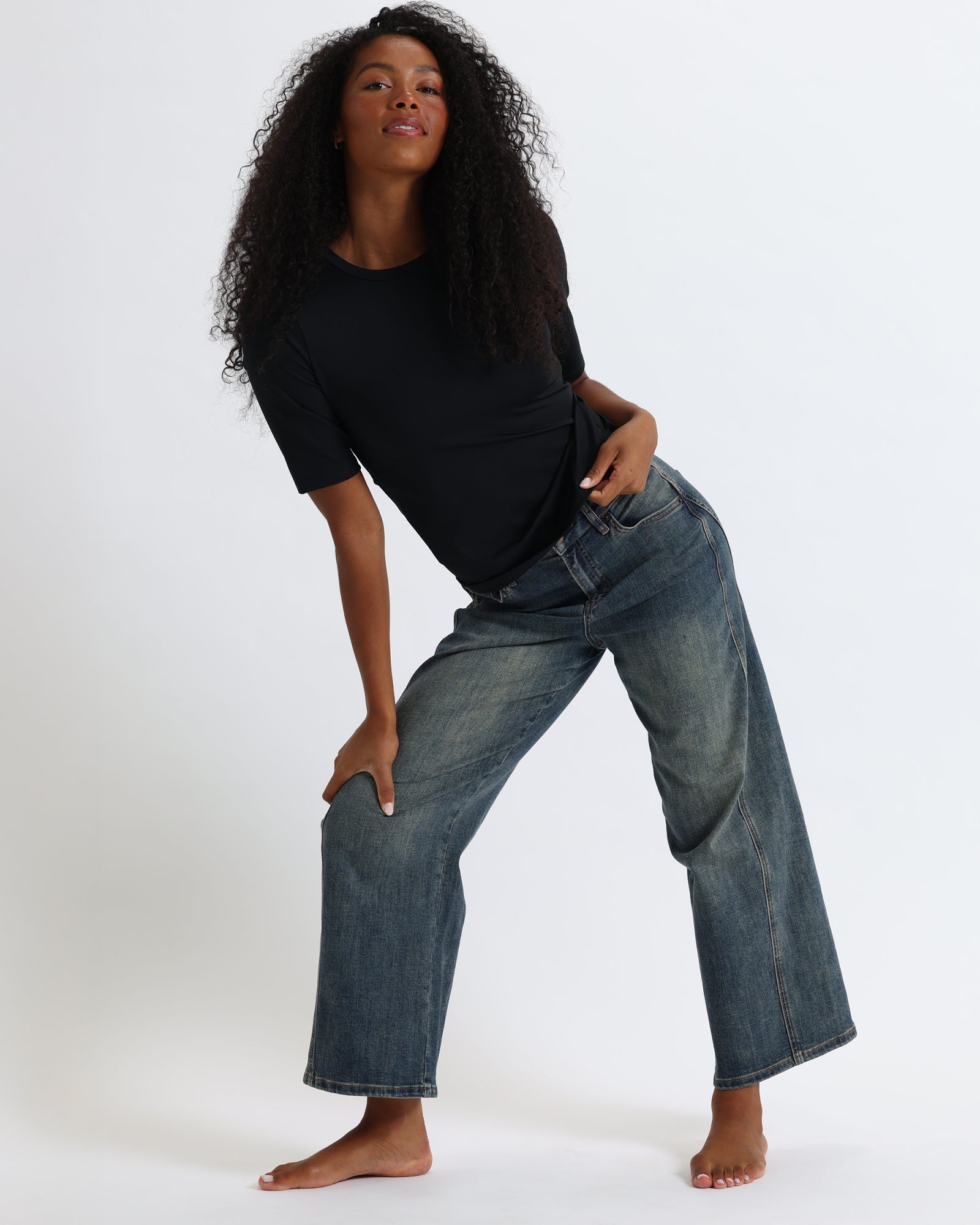 A woman with long, curly black hair is wearing a fitted black short-sleeve top and  jeans, standing against a white backdrop.
