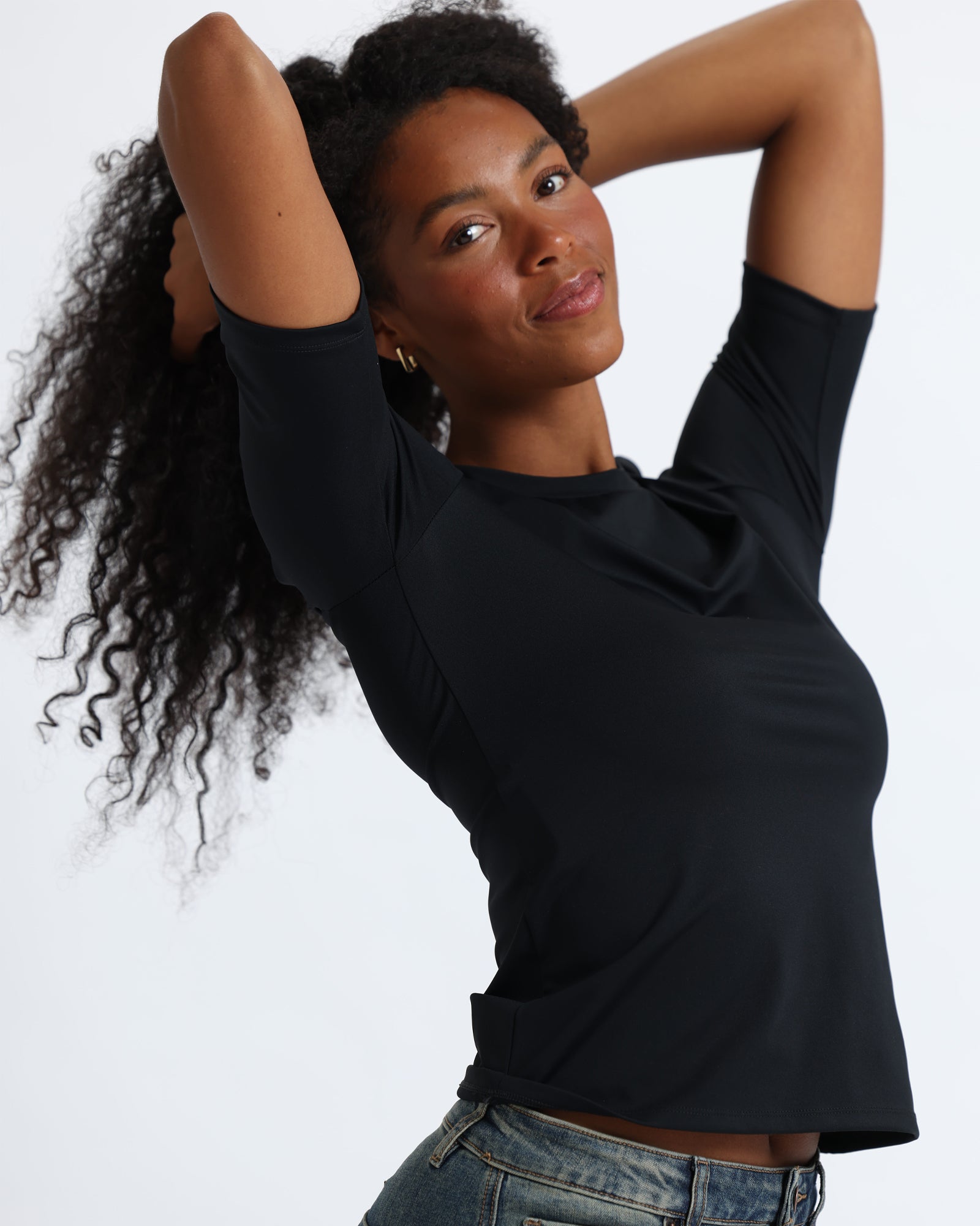 A woman with long, curly black hair is wearing a fitted black short-sleeve top and  jeans, standing against a white backdrop.