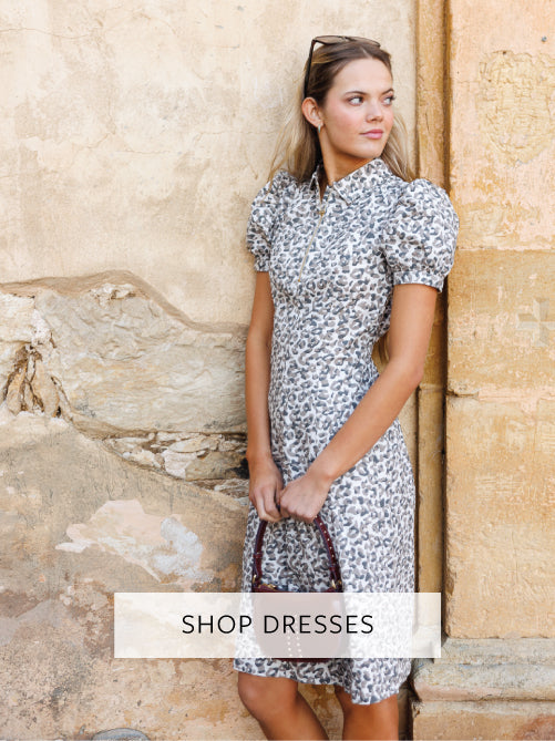 Woman wearing a patterned dress leaning against a stone wall with 'Shop Dresses' text overlay.