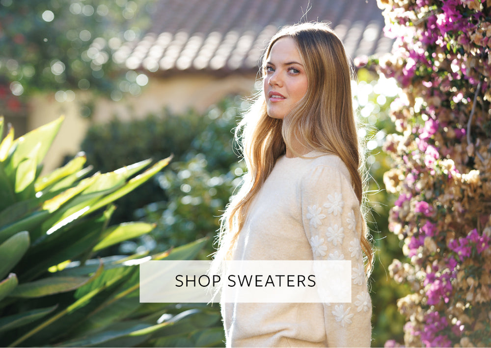 Woman in a garden wearing a sweater with text 'Shop Sweaters'.