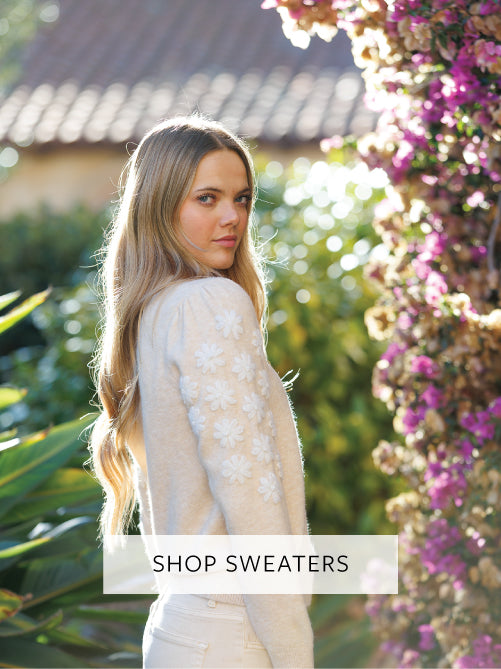 Woman in a garden wearing a sweater with text 'Shop Sweaters'.