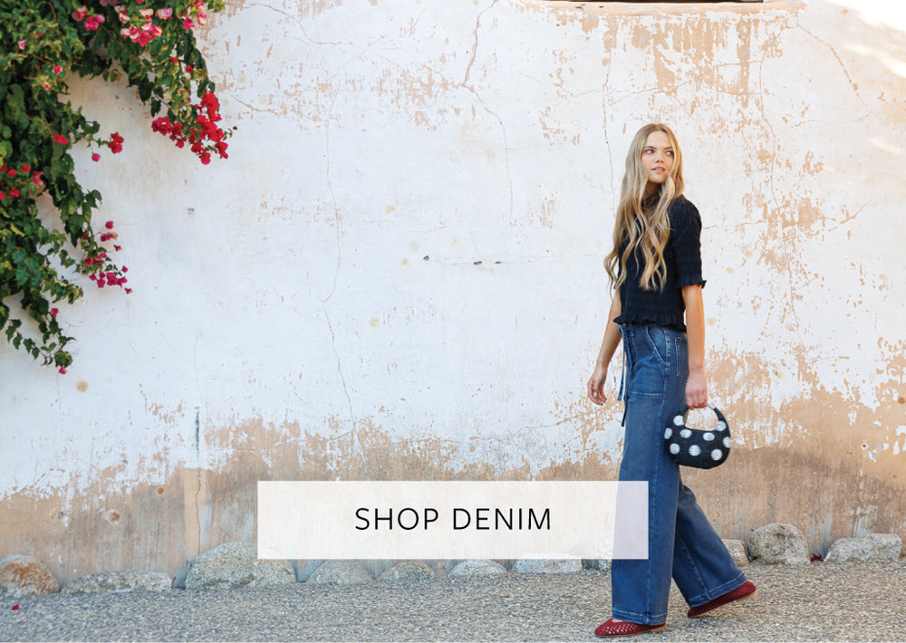 Woman in denim jeans standing against a textured wall with floral decorations, with 'Shop Denim' text overlay.