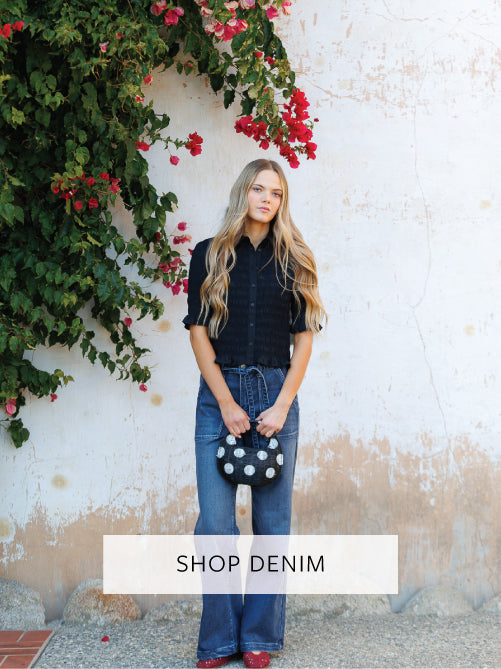 Woman in denim jeans standing against a textured wall with floral decorations, with 'Shop Denim' text overlay.