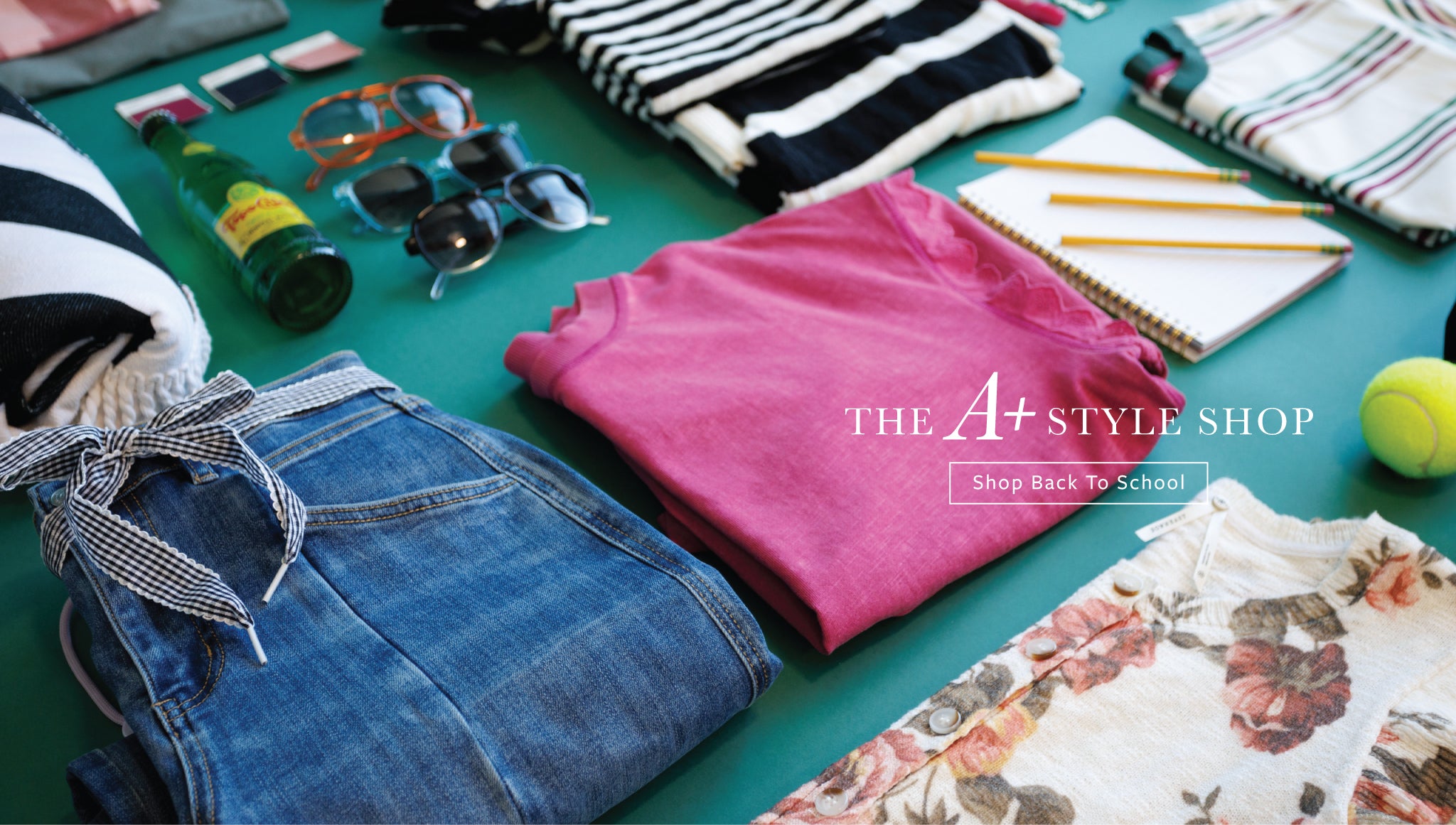 A styled flat-lay of folded clothing and accessories on a teal surface, including a bright pink T-shirt, blue jeans with a striped tie, striped knitwear, sunglasses, a notebook, pencils, a tennis ball, and floral fabric. The text 'THE A+ STYLE SHOP' is overlaid in the center.