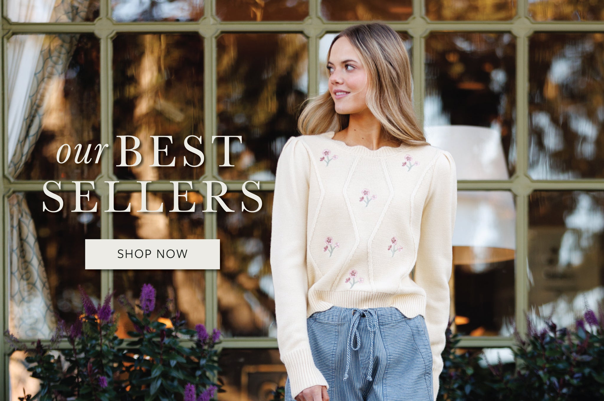 Woman wearing a beige sweater with floral embroidery in front of a window display.