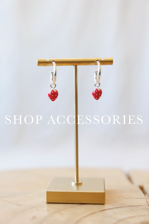 A pair of small red heart-shaped drop earrings with silver hoops, displayed on a gold stand over a wooden surface with a soft white background and the text 'SHOP ACCESSORIES' overlaid.