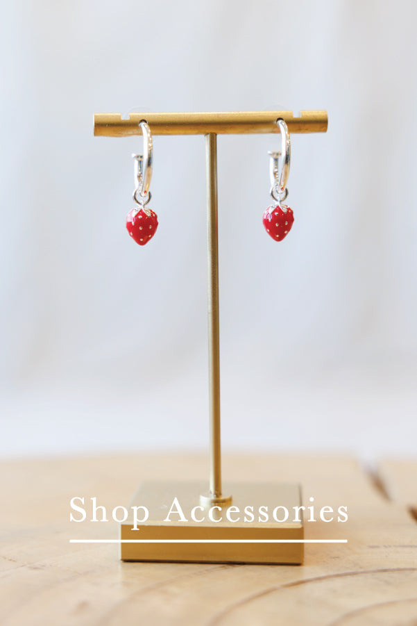 A pair of silver huggie hoop earrings with small red heart charms, displayed on a gold-colored T-stand. The background is a soft, out-of-focus light gray, with a wooden surface at the bottom.