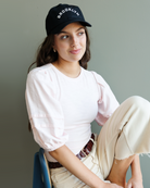 Woman wearing a black 'Brooklyn' cap and light pink shirt sitting on a chair against a gray background