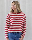 A woman wearing a red and off-white horizontally striped long-sleeve top with a wide boat neckline, paired with blue jeans, standing against a light neutral curtain background.