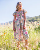A woman stands in a sunlit grassy field wearing a knee-length floral dress with short sleeves and a gathered waist. The dress has a beige base with pink, red, and green floral print.