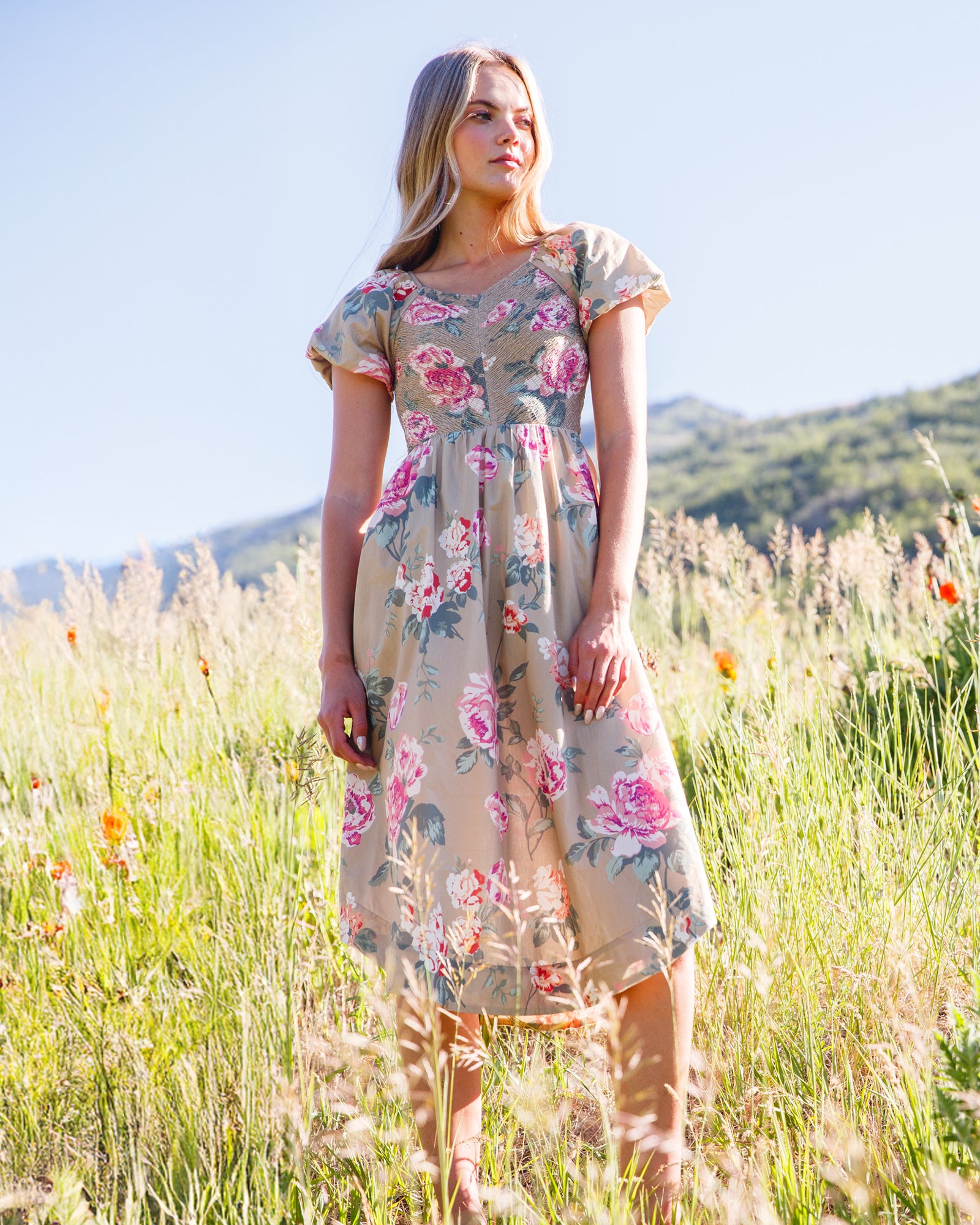 A woman stands in a sunlit grassy field wearing a knee-length floral dress with short sleeves and a gathered waist. The dress has a beige base with pink, red, and green floral print.