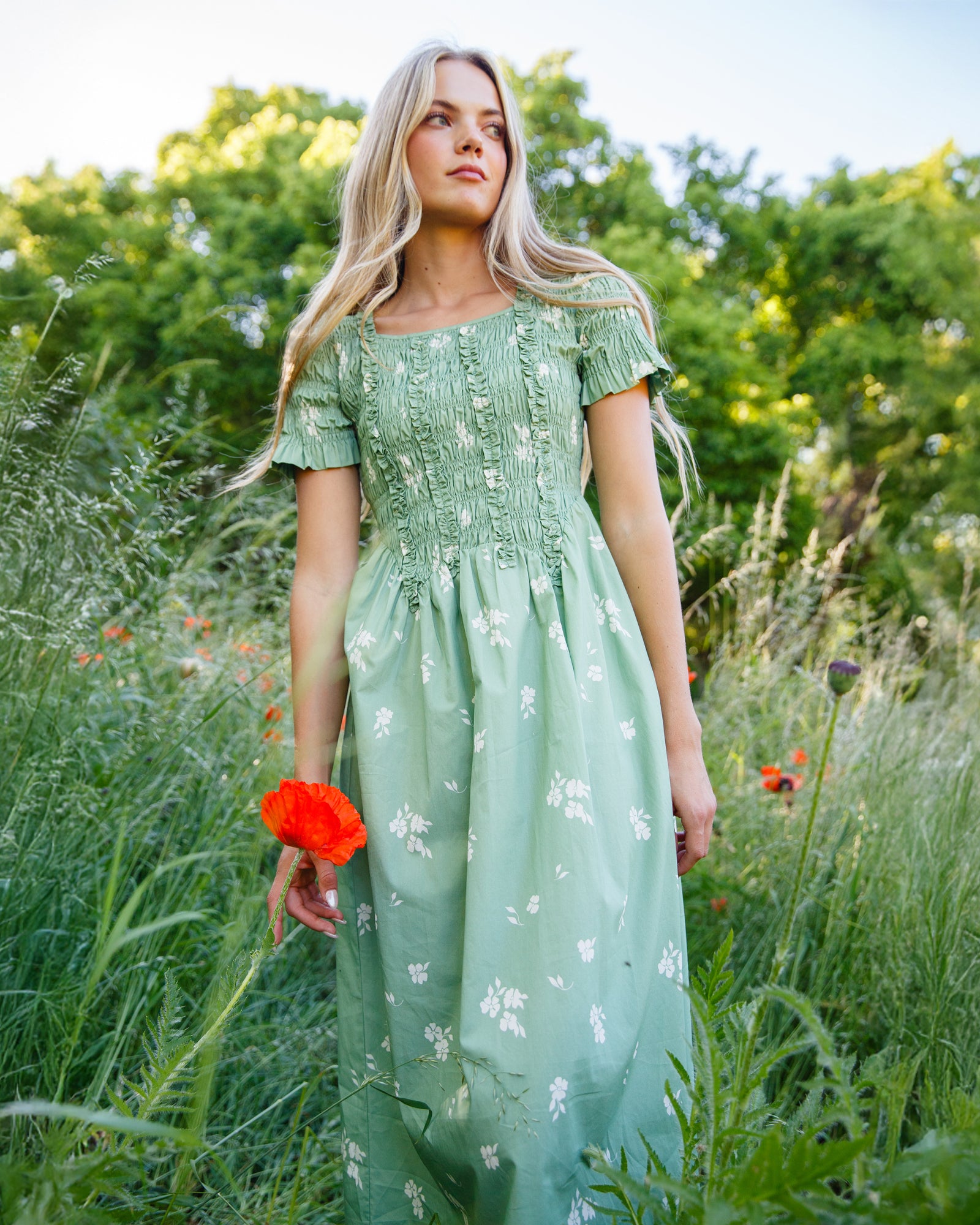A woman stands in a grassy outdoor field wearing a light sage green midi dress with white floral embroidery and short puff sleeves. She is holding a bright red poppy flower.