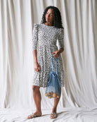 Woman in a patterned dress holding a blue bag against a white curtain background