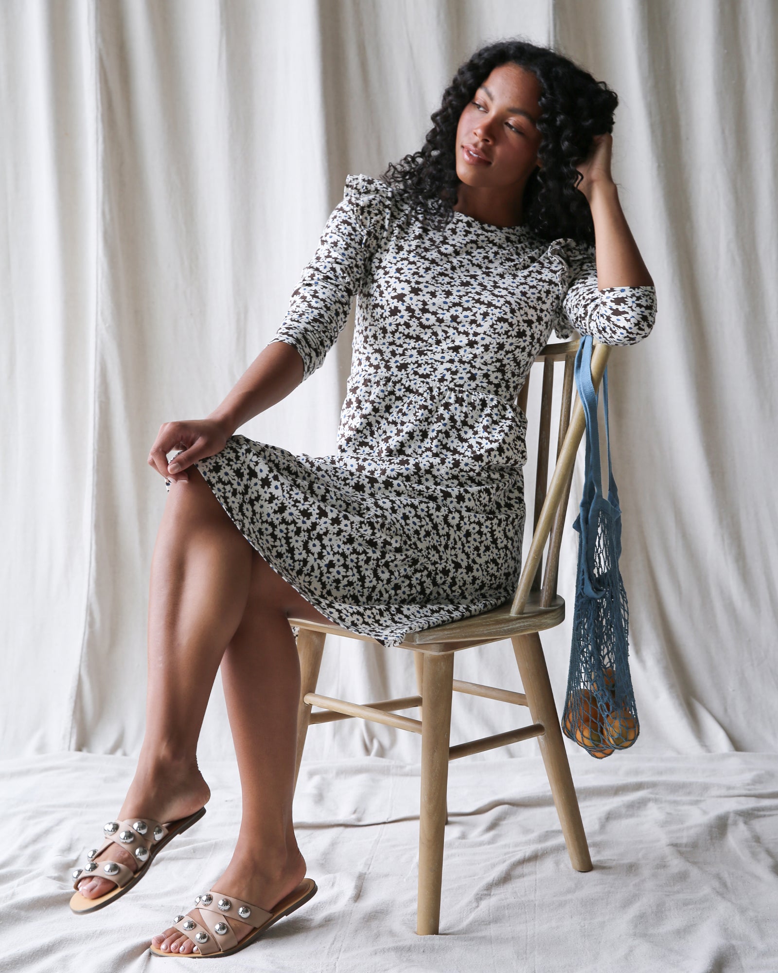 Woman sitting on a wooden chair wearing a black and white floral dress against a white curtain background.