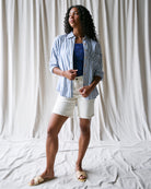 Woman wearing a blue striped shirt, navy top, white shorts, and sandals against a white curtain background.