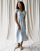 Woman wearing a light blue floral dress standing against a white curtain.