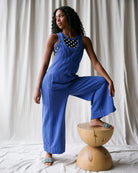 Woman in a blue jumpsuit sitting on a wooden stool against a white curtain background