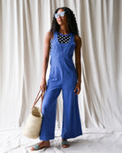 Woman wearing a blue jumpsuit with a polka dot top, holding a straw bag against a white curtain background.