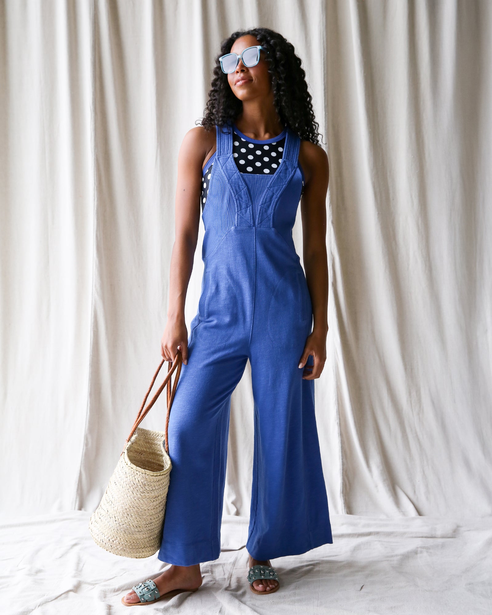 Woman wearing a blue jumpsuit with a polka dot top, holding a straw bag against a white curtain background.