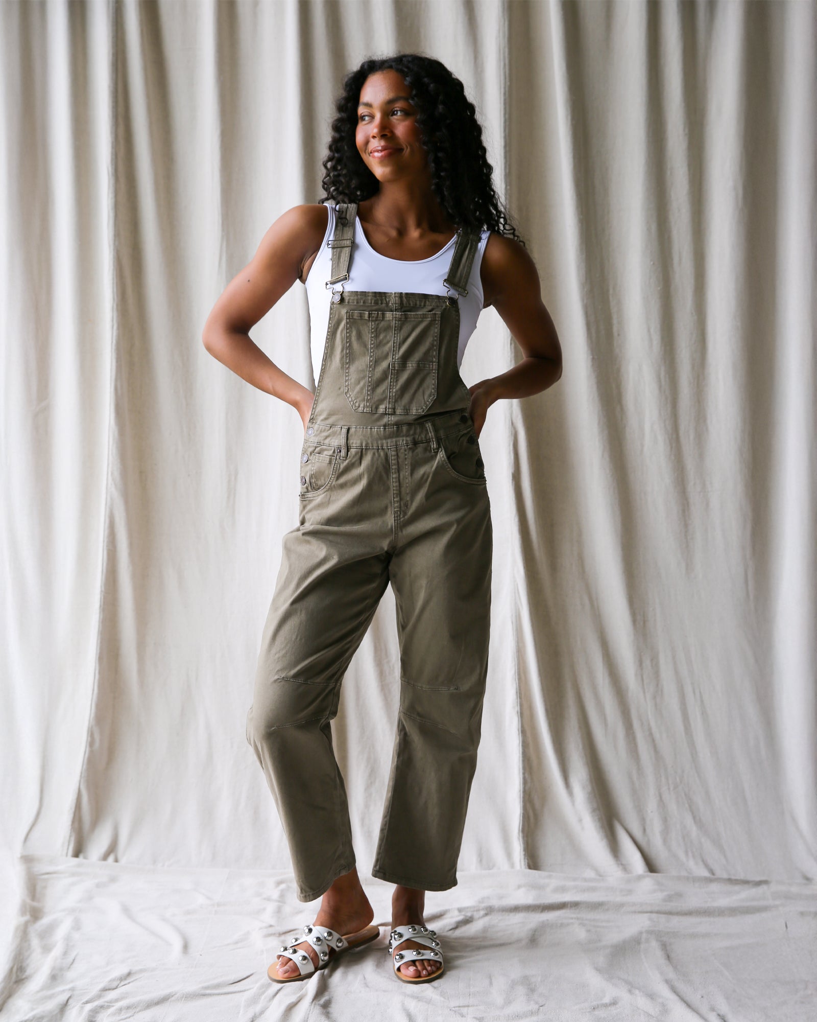 Woman wearing beige overalls and a white tank top against a plain background
