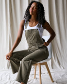 Woman wearing green overalls sitting on a chair against a white curtain background