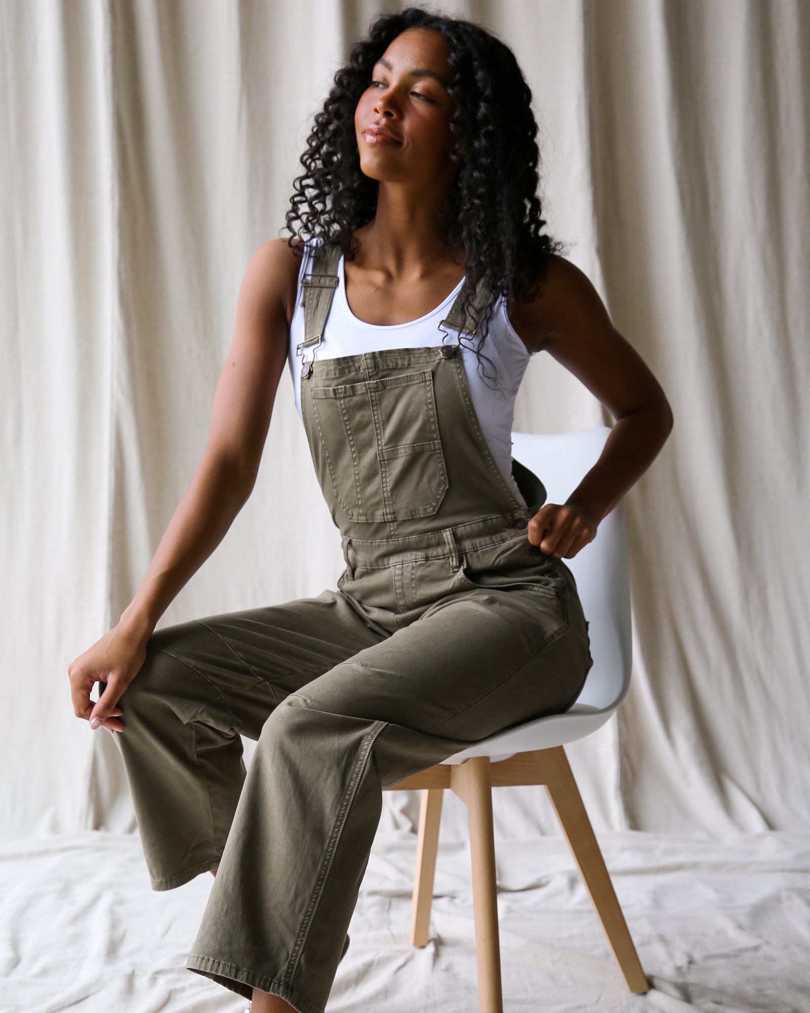 Woman wearing green overalls sitting on a chair against a white curtain background