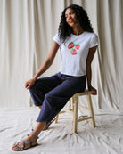 Woman sitting on a stool wearing a white t-shirt with strawberry design and navy pants against a neutral background.