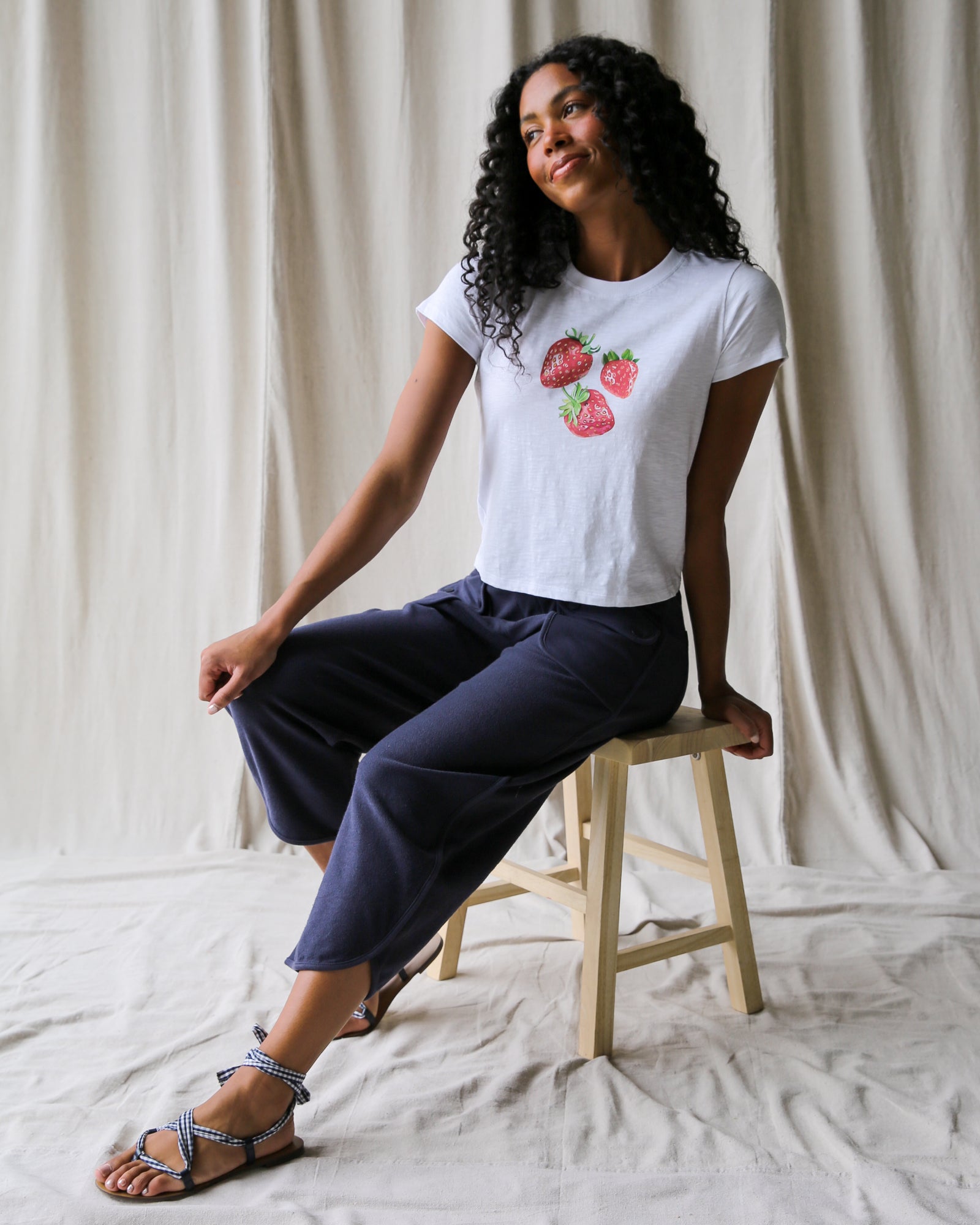 Woman sitting on a stool wearing a white t-shirt with strawberry design and navy pants against a neutral background.