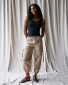 Woman wearing a black tank top and beige cargo pants standing against a white curtain.