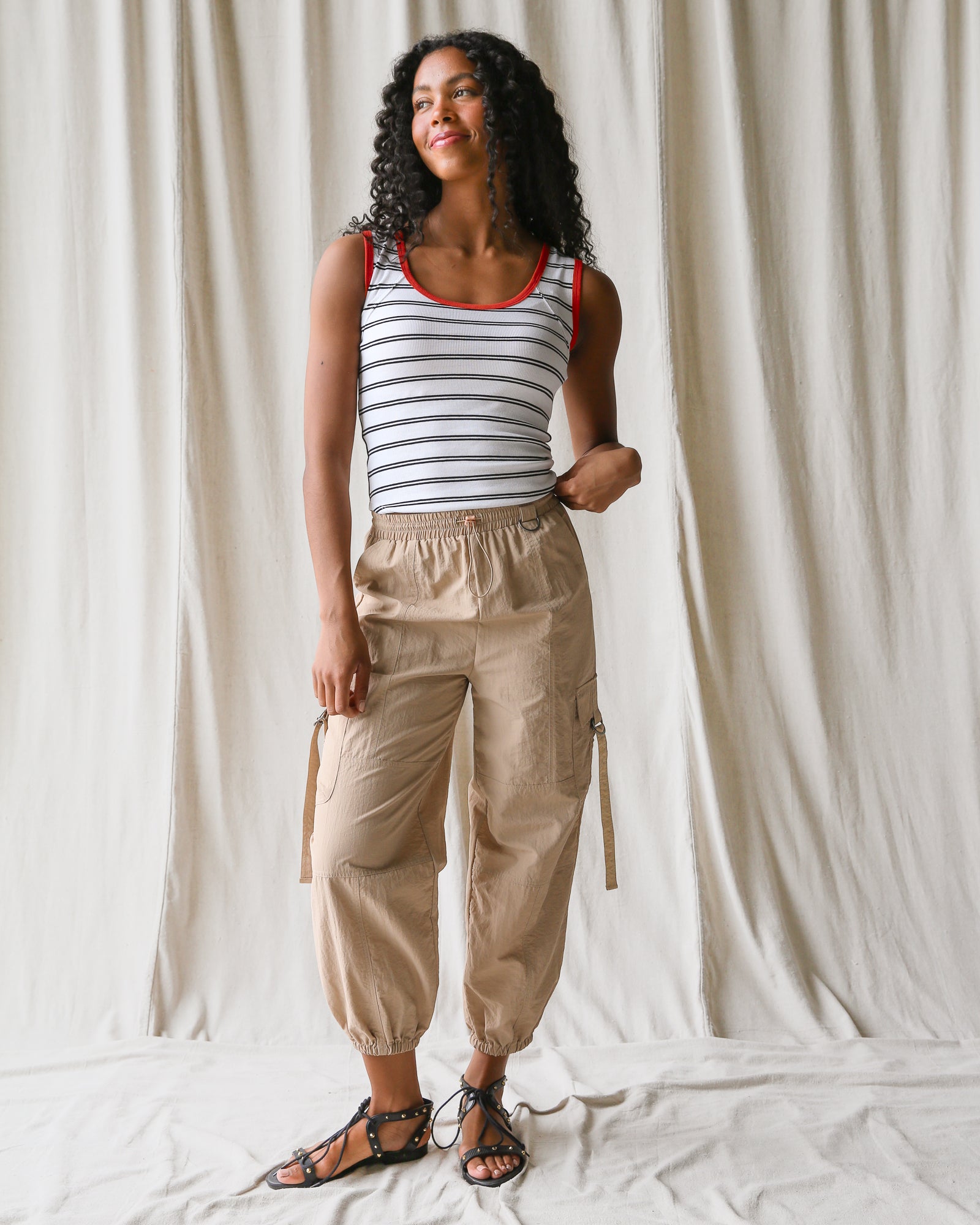 Woman wearing a striped tank top and beige cargo pants against a white curtain background