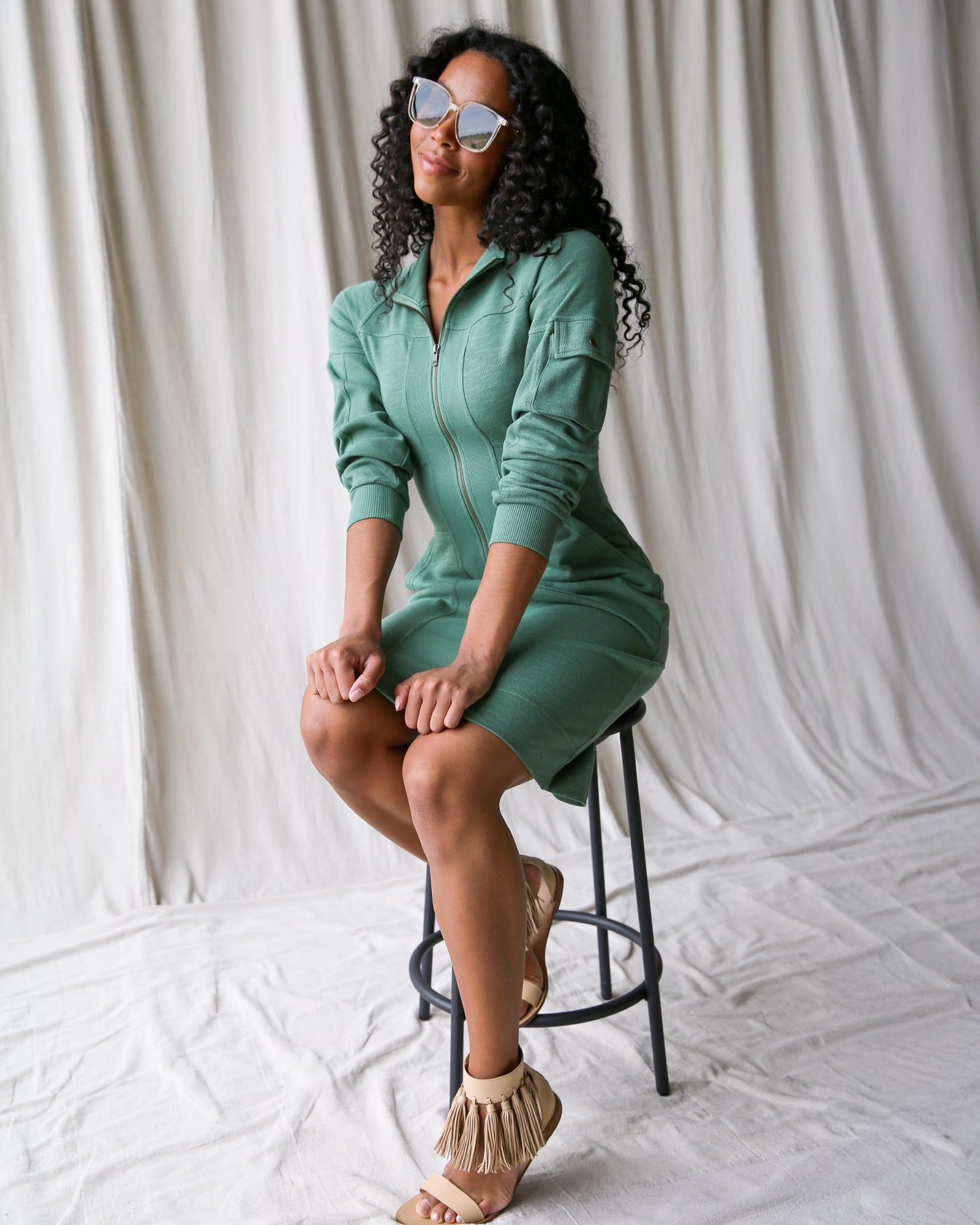 Woman in a green dress sitting on a stool against a white curtain background
