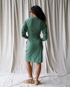 Woman wearing a green dress standing against a plain white curtain.