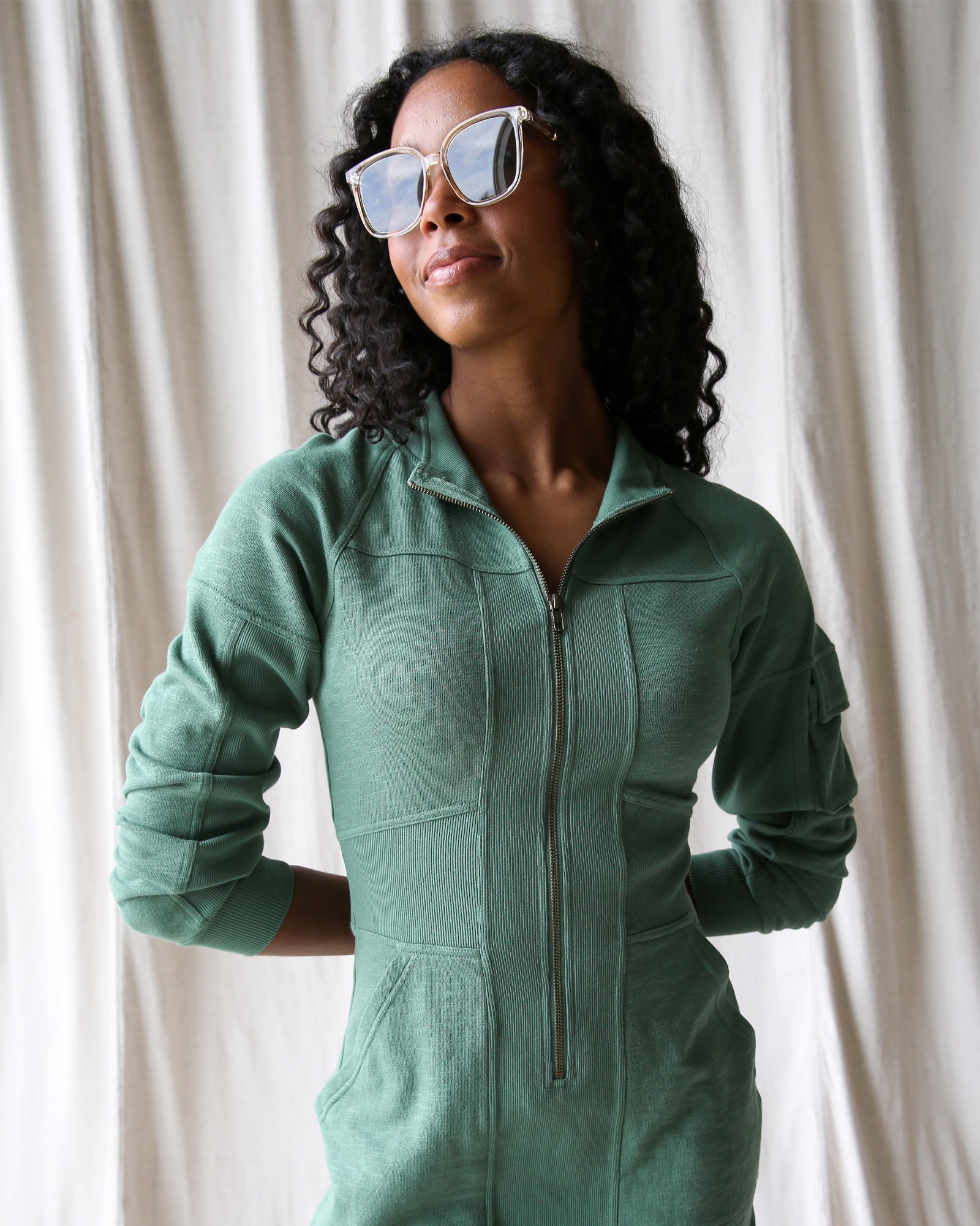 Woman wearing a green onesie against a white curtain background