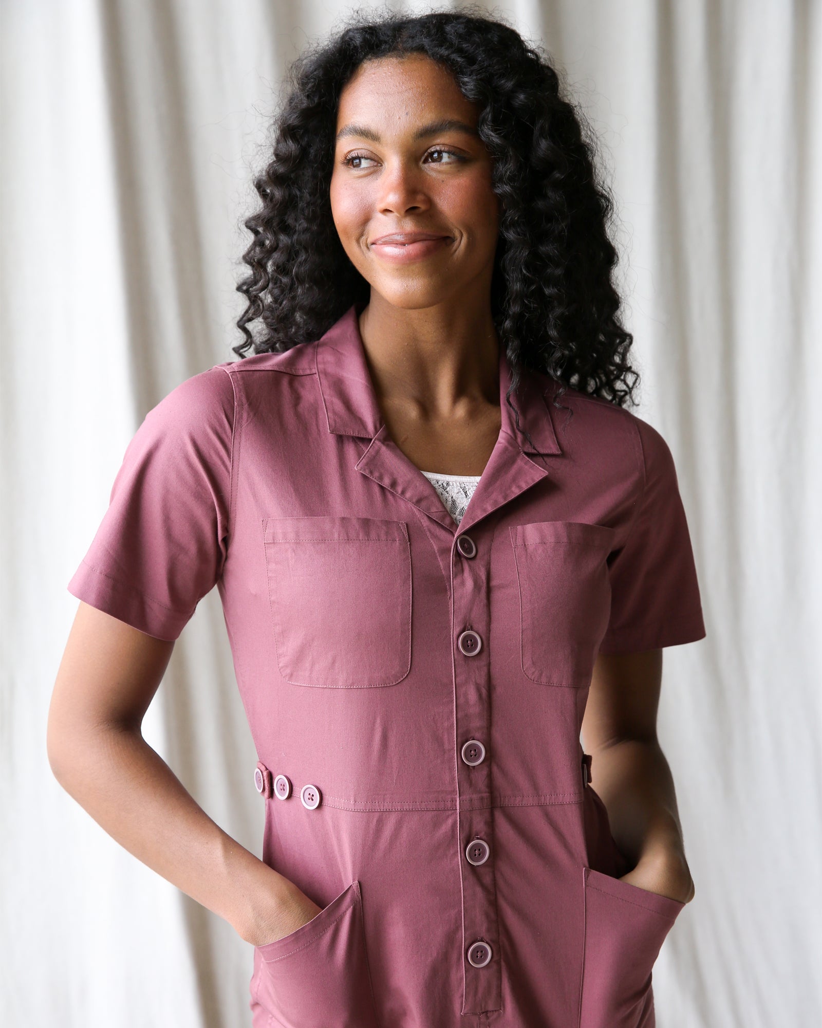 Woman wearing a pink jumpsuit against a white curtain background