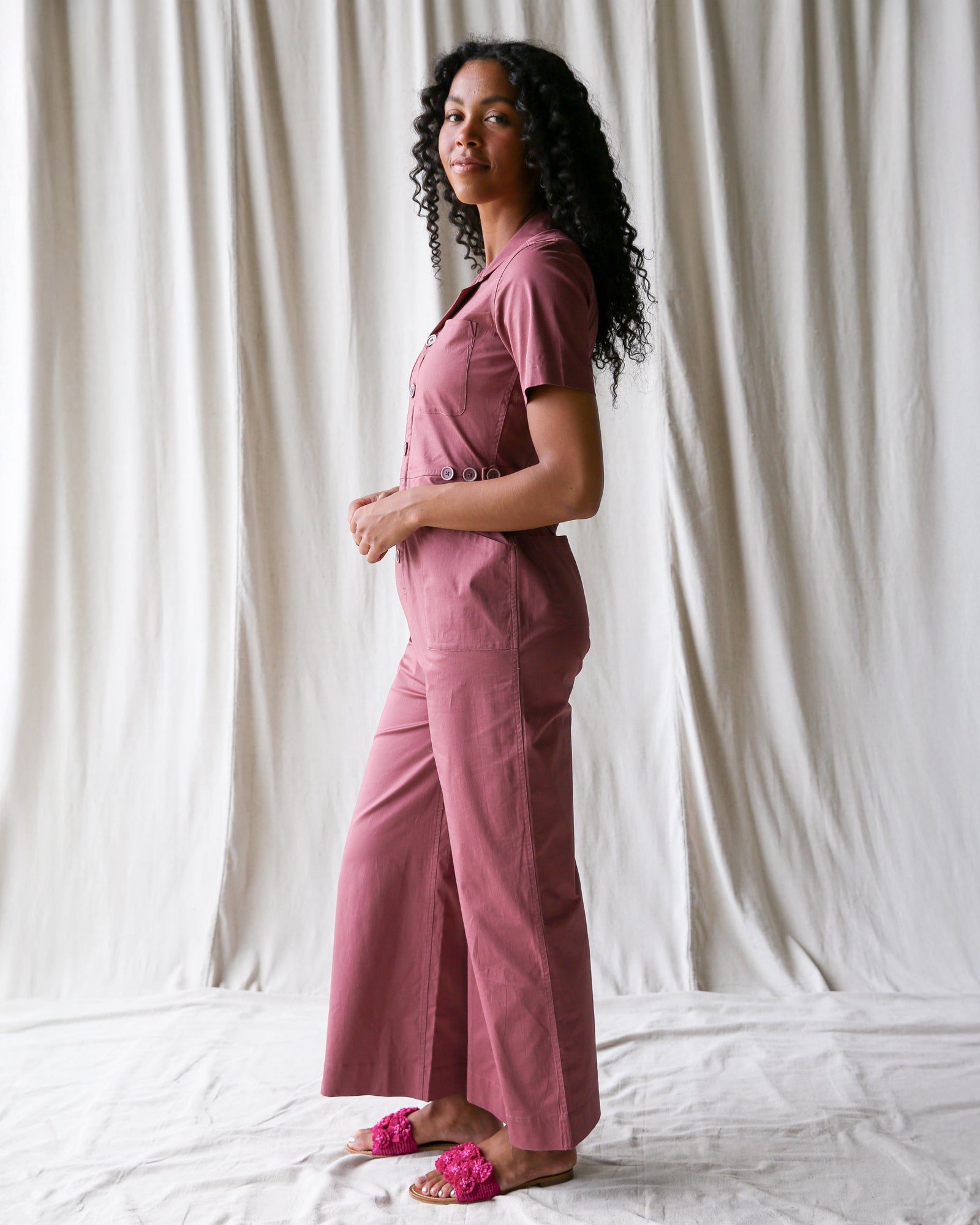 Woman wearing a pink jumpsuit standing against a white curtain.