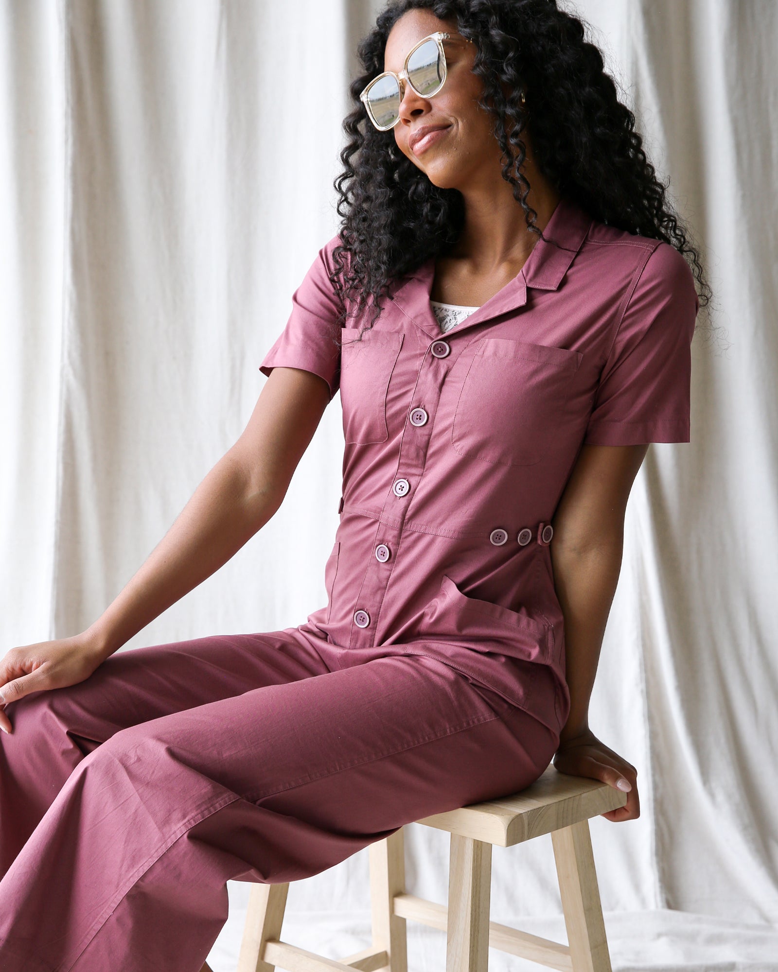 Woman wearing a pink jumpsuit sitting on a stool against a white curtain background