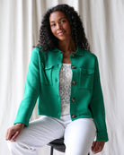 Woman wearing a green jacket over a white top with a pattern, sitting against a white curtain background.