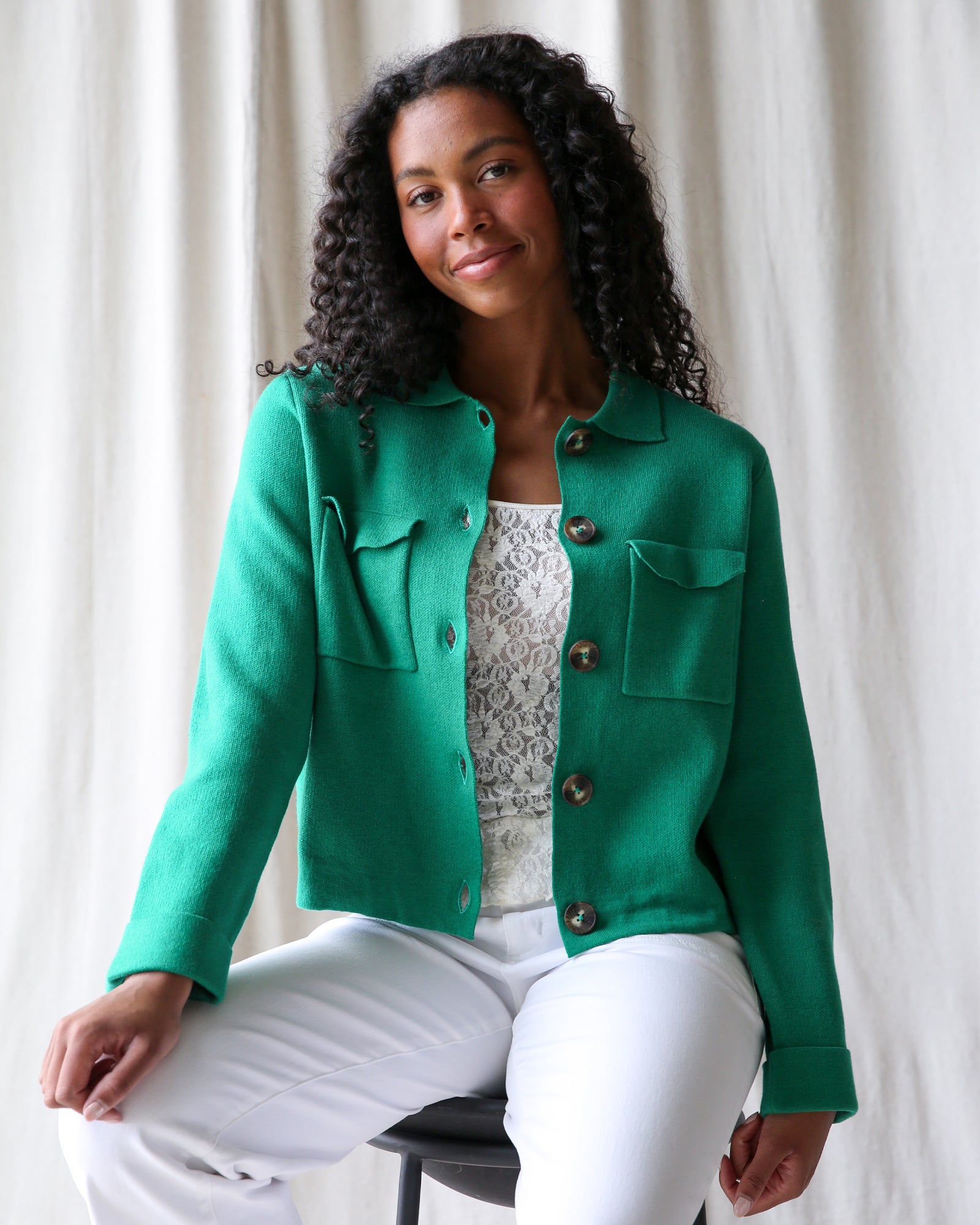 Woman wearing a green jacket over a white top with a pattern, sitting against a white curtain background.