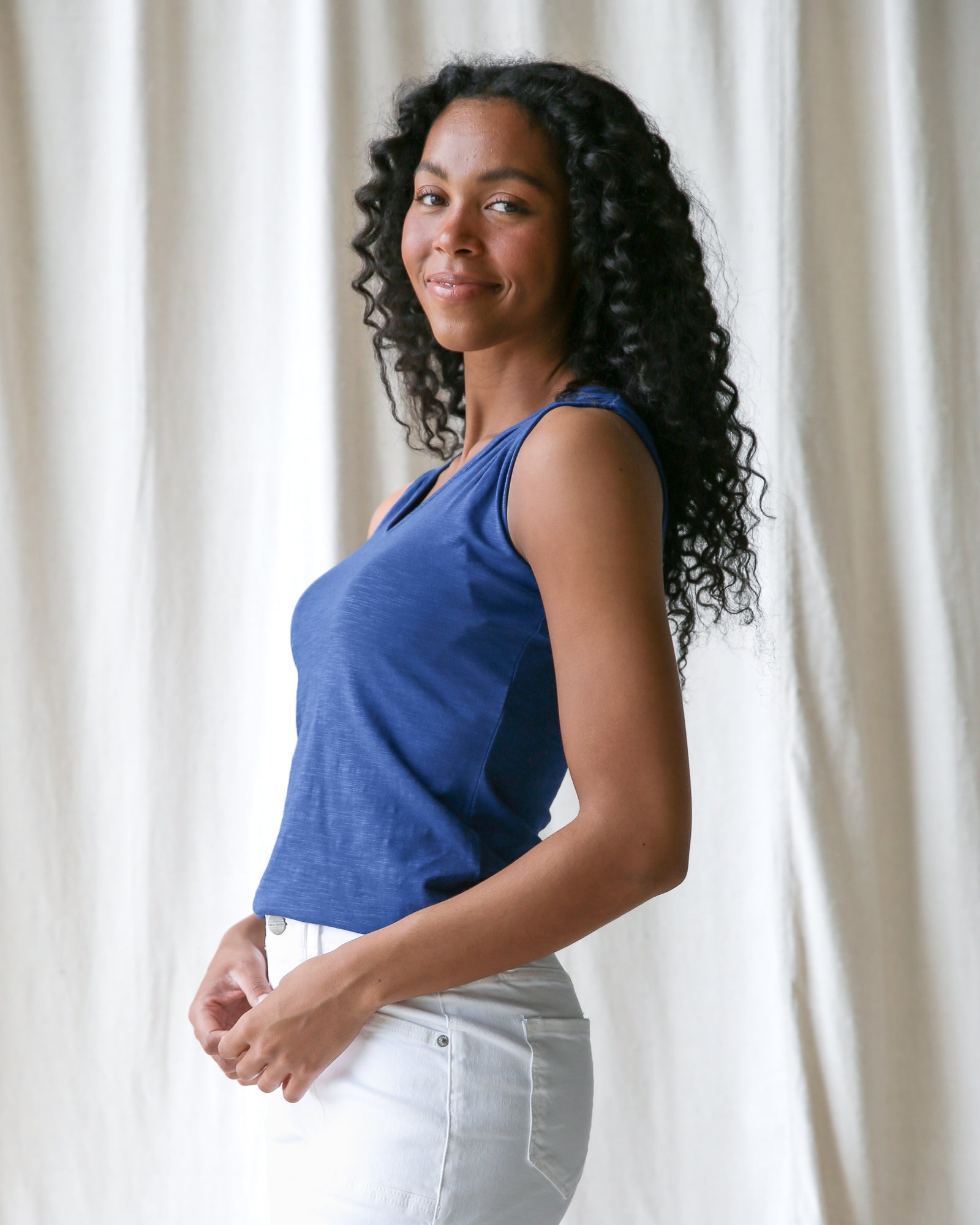 Woman wearing a blue top and white pants standing against a white curtain.