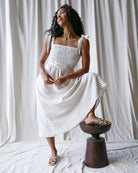 Woman in a white dress sitting on a wooden stool against a white curtain background