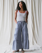 Woman wearing a striped tank top and blue tiered pants against a white curtain background