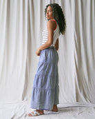 Woman wearing a striped top and tiered blue skirt standing against a plain white curtain.