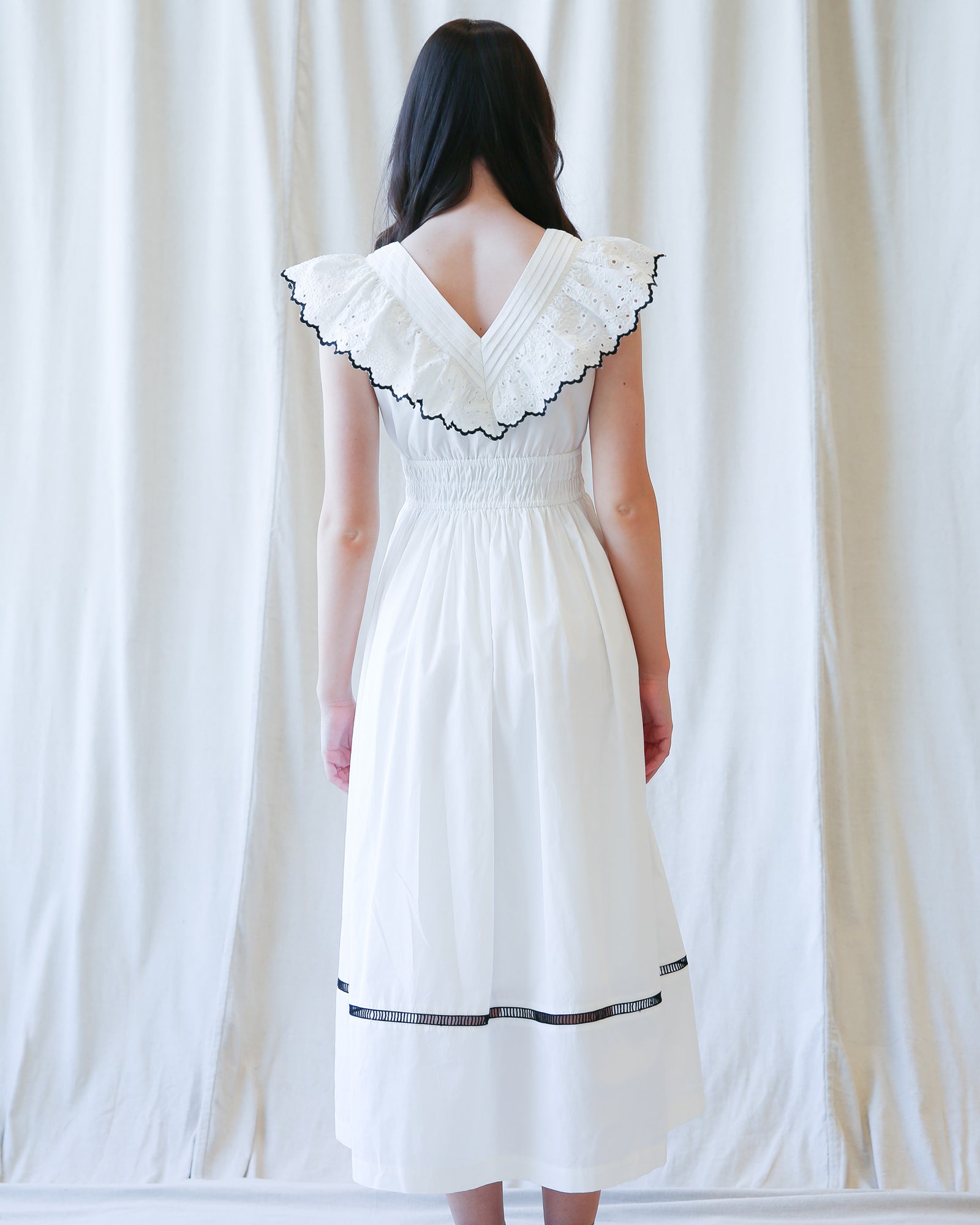 Woman wearing a white dress with black trim against a white curtain background