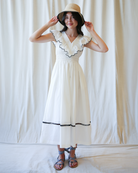 Woman wearing a white dress with black trim and a straw hat against a plain background