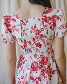 Close up Image of the back of a woman wearing a white dress with red floral print standing in front of a white canvas background