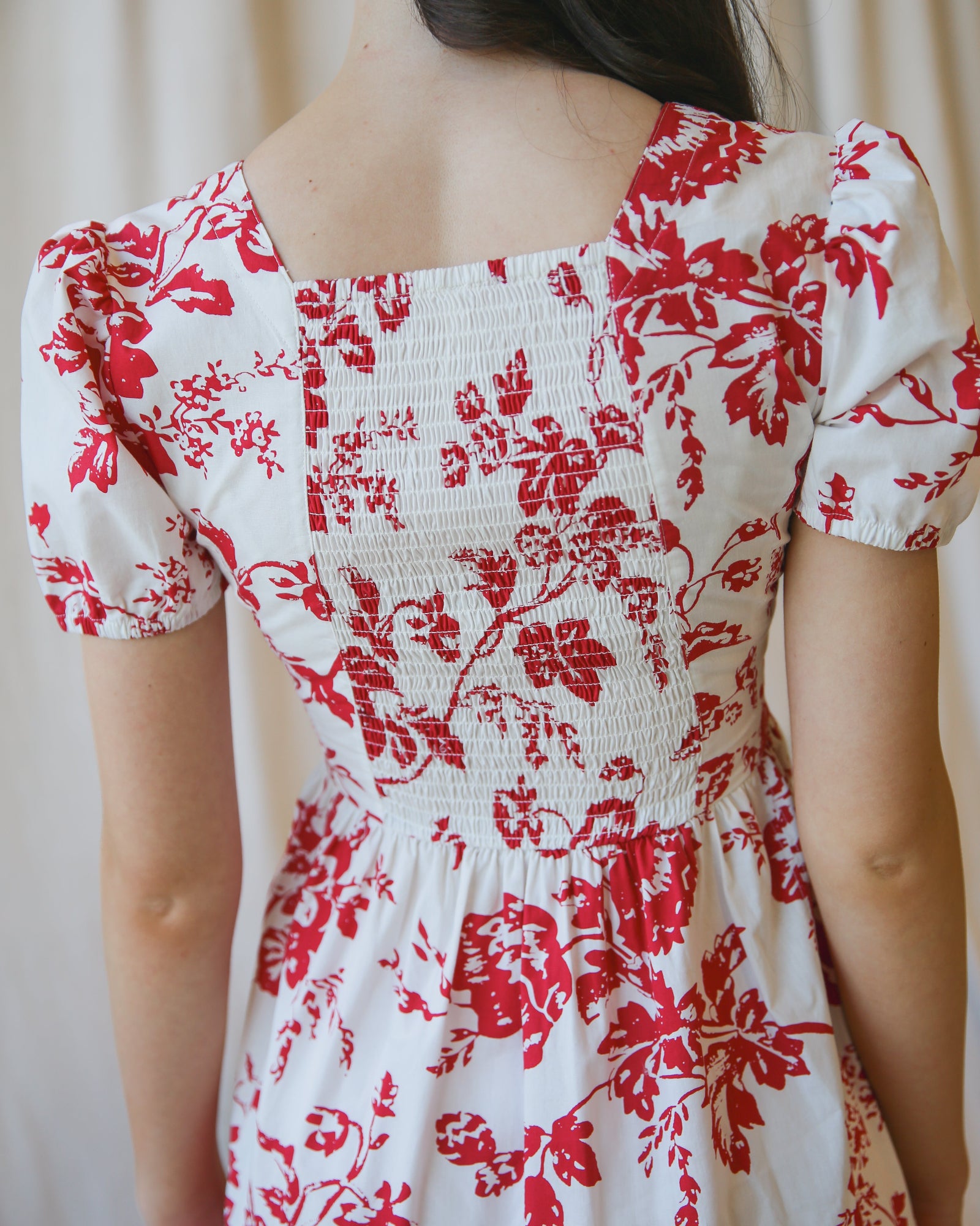 Close up Image of the back of a woman wearing a white dress with red floral print standing in front of a white canvas background